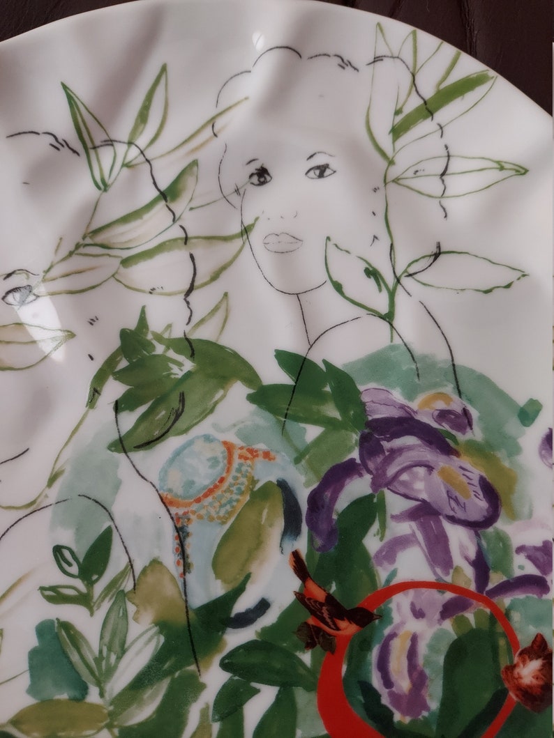 Puede incluir: Un plato de cer&aacute;mica blanco con un dise&ntilde;o floral estilizado que presenta un rostro de mujer, hojas verdes y flores moradas. El plato tiene un borde ondulado.