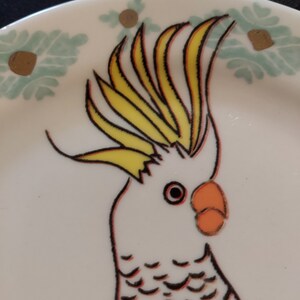 May include: A white cockatoo with a yellow crest and orange beak is depicted on a white ceramic plate with a green and gold floral design.