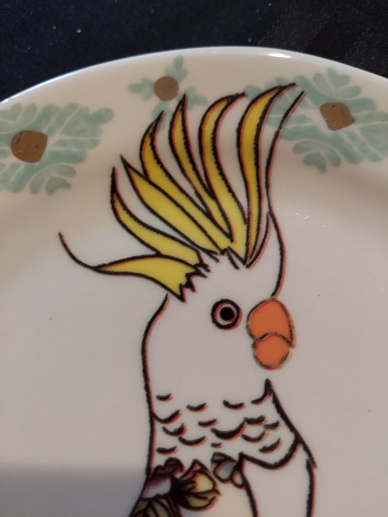 May include: A white cockatoo with a yellow crest and orange beak is depicted on a white ceramic plate with a green and gold floral design.