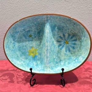 May include: An oval-shaped, divided serving dish with a turquoise interior and a brown rim. The dish features painted yellow and blue flowers. The dish is displayed on a black metal stand, set against a pink tablecloth.