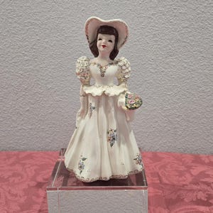 May include: A ceramic figurine of a woman in a white dress and hat, holding a floral basket. The dress is adorned with floral patterns and gold trim. The figurine is displayed on a clear, rectangular stand.