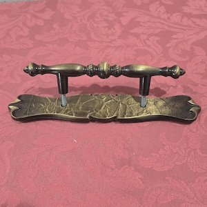 May include: A gold-toned metal drawer pull with a decorative, ornate design. The pull has a curved, rectangular base and a center bar with a decorative knob on each end.