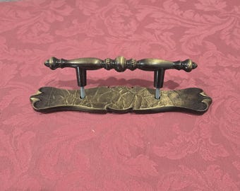 May include: A gold-toned metal drawer pull with a decorative, ornate design. The pull has a curved, rectangular base and a centre bar with a decorative knob on each end.