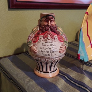 May include: A ceramic beer stein with a whimsical design of a bearded man wearing a crown and holding a beer mug. The stein is decorated with intricate details and the text "Reiner Wein und gutes Bier Sind des Wirtes schönste Zier".