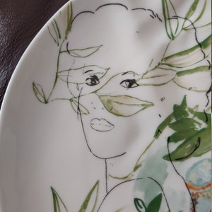 Puede incluir: Un plato de cer&aacute;mica blanco con un dibujo en l&iacute;nea negro y verde de la cara de una mujer rodeada de hojas y flores.