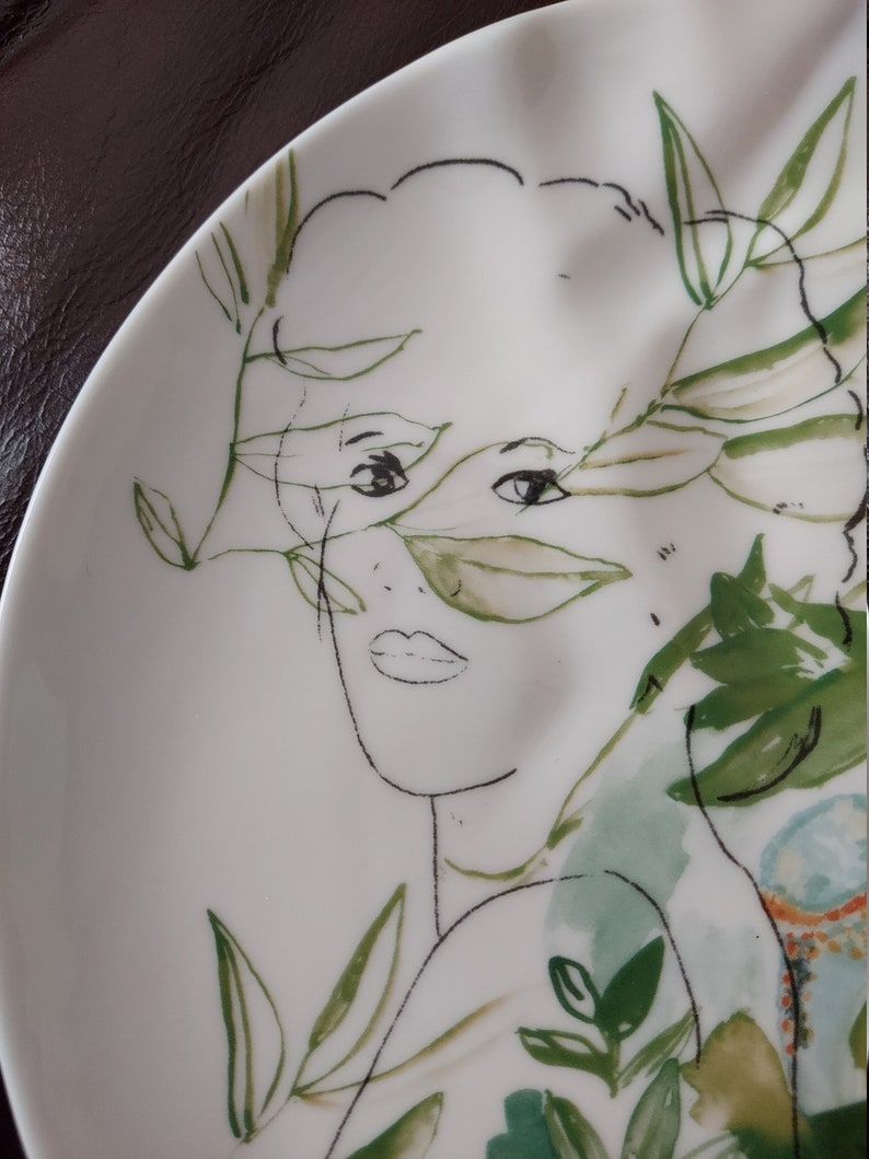 Puede incluir: Un plato de cer&aacute;mica blanco con un dibujo en l&iacute;nea negro y verde de la cara de una mujer rodeada de hojas y flores.