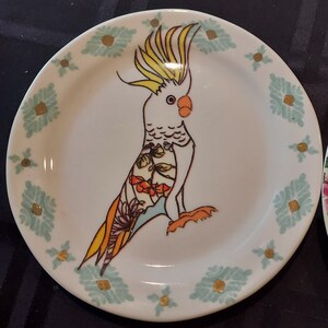 May include: A white ceramic plate with a blue and gold floral design. A hand-painted cockatoo with yellow and orange feathers is on the plate.