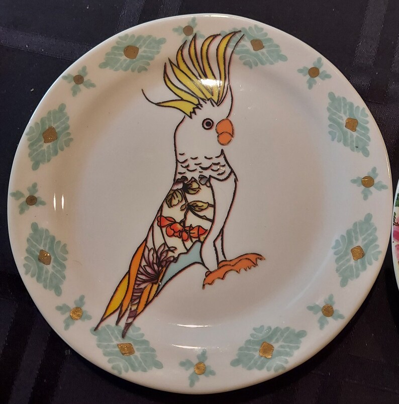 May include: A white ceramic plate with a blue and gold floral design. A hand-painted cockatoo with yellow and orange feathers is on the plate.