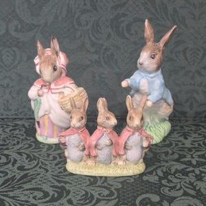 May include: A collection of five ceramic rabbit figurines. One rabbit wears a pink bonnet and cape with a basket, another wears a blue jacket, and three smaller rabbits stand together on a grassy base, each wearing a small pink cape.