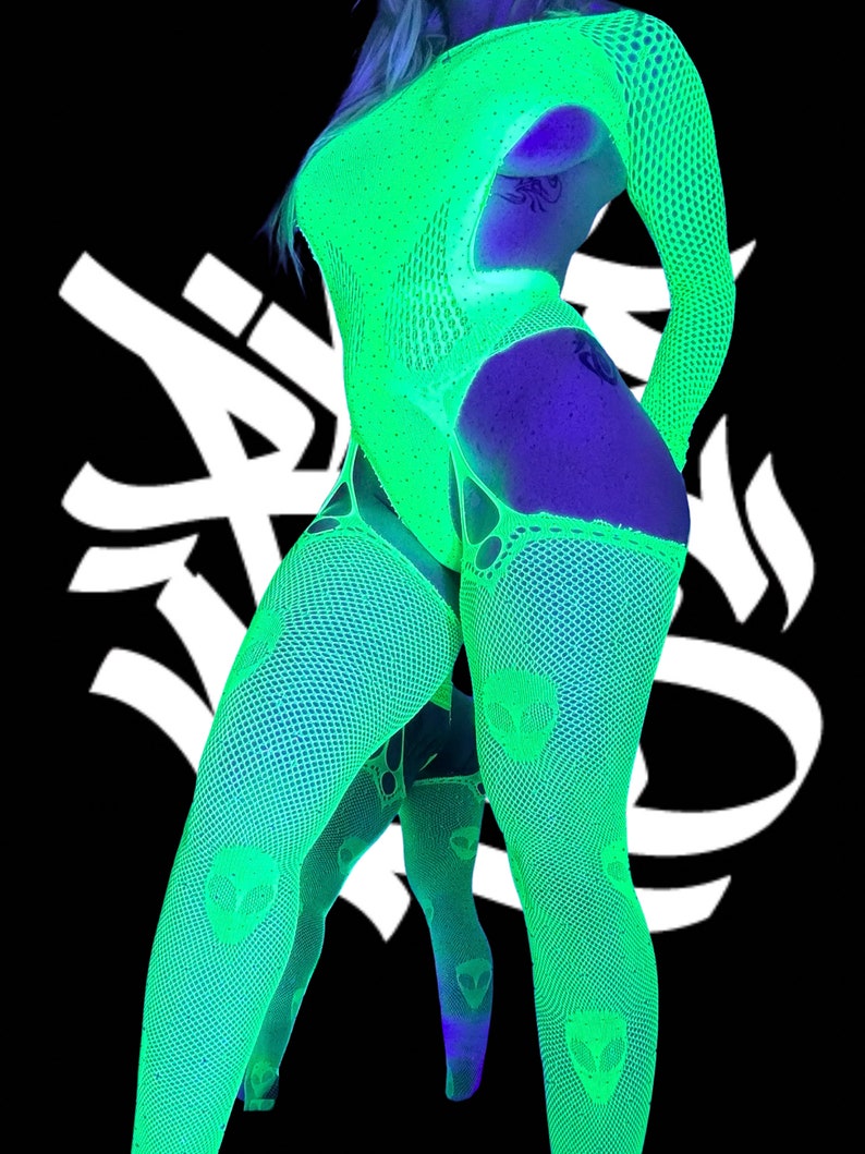 May include: A woman wearing a green and black fishnet bodysuit with alien head graphics. The bodysuit has a plunging neckline and long sleeves. The woman is standing with her legs crossed.