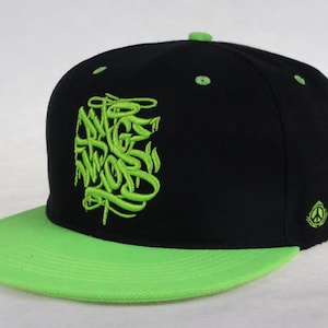 Puede incluir: Una gorra de béisbol negra y verde con un diseño bordado en verde en la parte delantera. La gorra tiene una visera verde y una corona negra.
