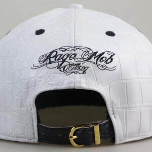 May include: White leather baseball cap with a black crocodile pattern and black embroidery that reads "Rage Mob Clothing". The cap has a black strap with a gold buckle.
