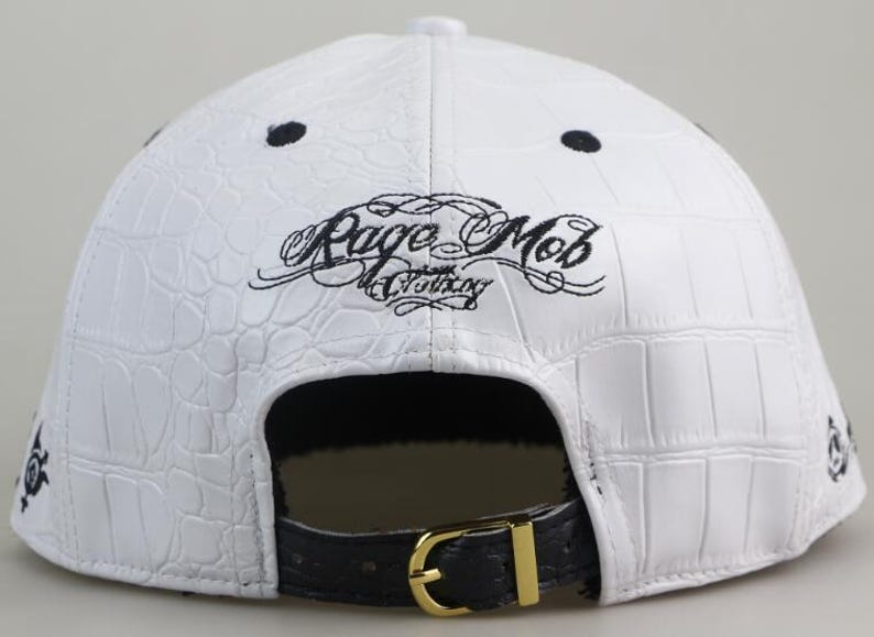 May include: White leather baseball cap with a black crocodile pattern and black embroidery that reads "Rage Mob Clothing". The cap has a black strap with a gold buckle.