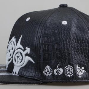 Puede incluir: Gorra de b&eacute;isbol de cuero negro con dise&ntilde;os bordados en blanco. La gorra presenta un patr&oacute;n de piel de cocodrilo y una visera plana con rayas blancas.