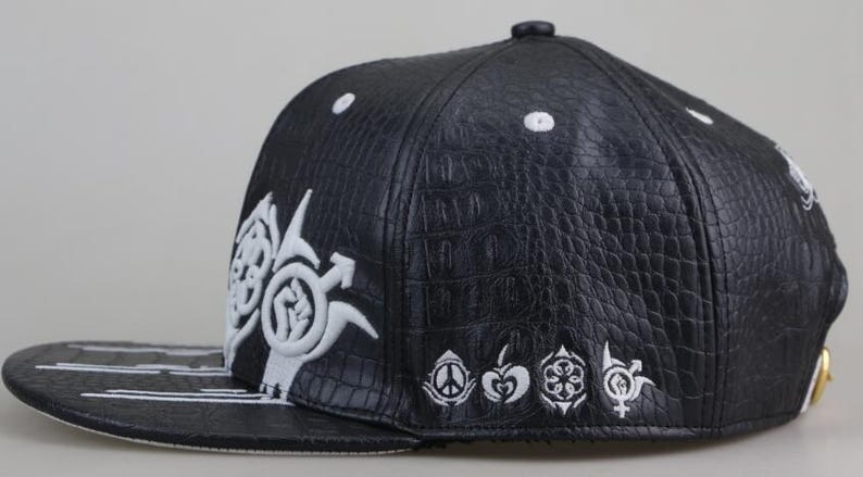 Puede incluir: Gorra de b&eacute;isbol de cuero negro con dise&ntilde;os bordados en blanco. La gorra presenta un patr&oacute;n de piel de cocodrilo y una visera plana con rayas blancas.