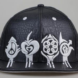 Puede incluir: Gorra de b&eacute;isbol de cuero negro con textura de cocodrilo. La gorra presenta s&iacute;mbolos bordados en blanco, incluyendo un signo de paz, un coraz&oacute;n, una flor y un dise&ntilde;o estilizado.