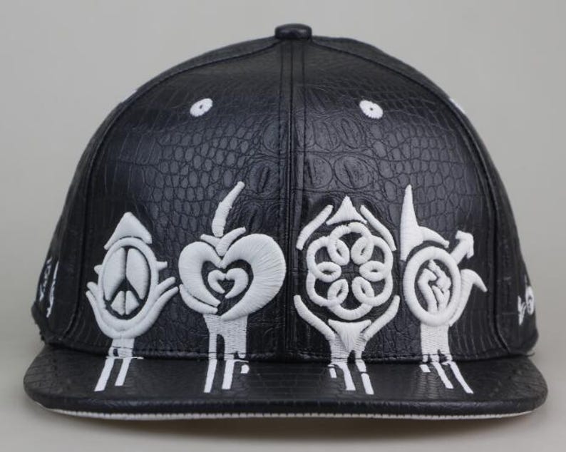 Puede incluir: Gorra de b&eacute;isbol de cuero negro con textura de cocodrilo. La gorra presenta s&iacute;mbolos bordados en blanco, incluyendo un signo de paz, un coraz&oacute;n, una flor y un dise&ntilde;o estilizado.
