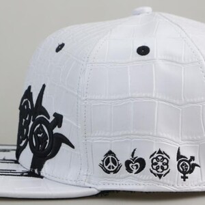 May include: White baseball cap with a black crocodile-embossed design. The cap features a black graphic on the front and a row of black symbols on the side.