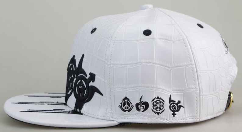 May include: White baseball cap with a black crocodile-embossed design. The cap features a black graphic on the front and a row of black symbols on the side.