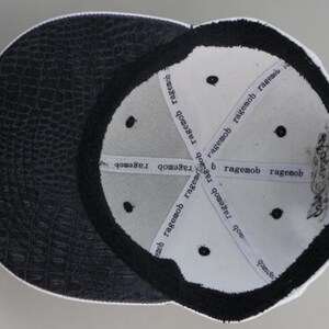 May include: White baseball cap with a black crocodile-embossed leather brim. The inside of the cap is white with black stitching and the words "ragemob" repeated around the inside.