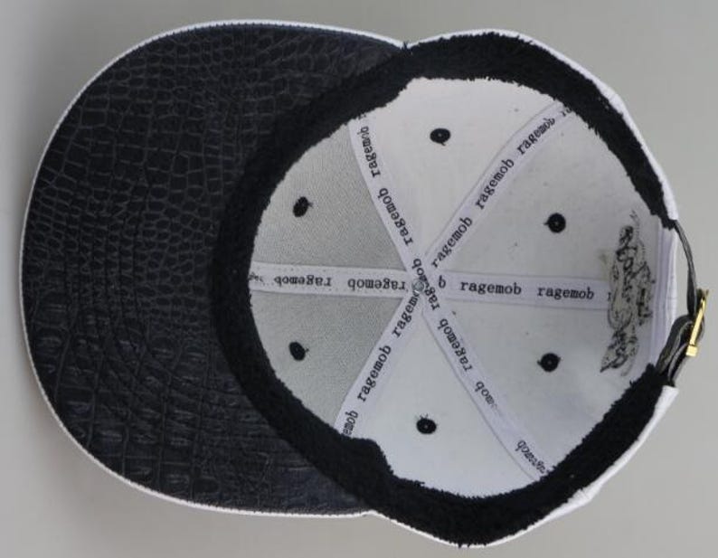 May include: White baseball cap with a black crocodile-embossed leather brim. The inside of the cap is white with black stitching and the words "ragemob" repeated around the inside.
