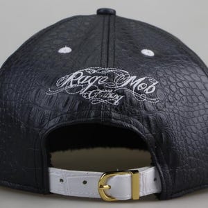 Puede incluir: Gorra de b&eacute;isbol de cuero negro con una correa de cuero blanco y un logotipo bordado en blanco que dice "Rage Mob Clothing".