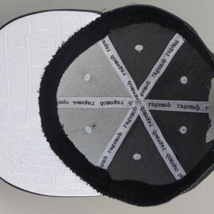 Puede incluir: Gorra de b&eacute;isbol negra y blanca con una visera de cuero blanco y una corona negra. El interior de la corona est&aacute; forrado con forro polar negro y tiene el texto "ragemob" repetido varias veces.