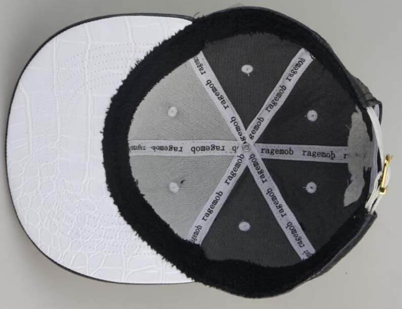 Puede incluir: Gorra de b&eacute;isbol negra y blanca con una visera de cuero blanco y una corona negra. El interior de la corona est&aacute; forrado con forro polar negro y tiene el texto "ragemob" repetido varias veces.