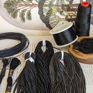 May include: A collection of black threads and cords for crafts. The image shows spools of thread, bundles of elastic cord, and skeins of embroidery floss. A small wooden bowl holds beaded cord. A botanical illustration is in the background.