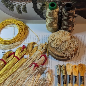 May include: A collection of gold-toned craft materials, such as metallic thread spools, wire bundles, and embroidery floss. These items are displayed on a printed paper surface, indicating a crafting or jewellery-making project.
