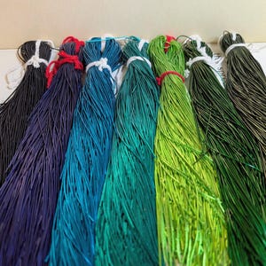 May include: Seven bundles of thin, flexible cord arranged in a row, featuring a gradient of colours from black, deep purple, and bright blue to teal, lime green, forest green, and olive green, each tied at the top with a small white or red string.