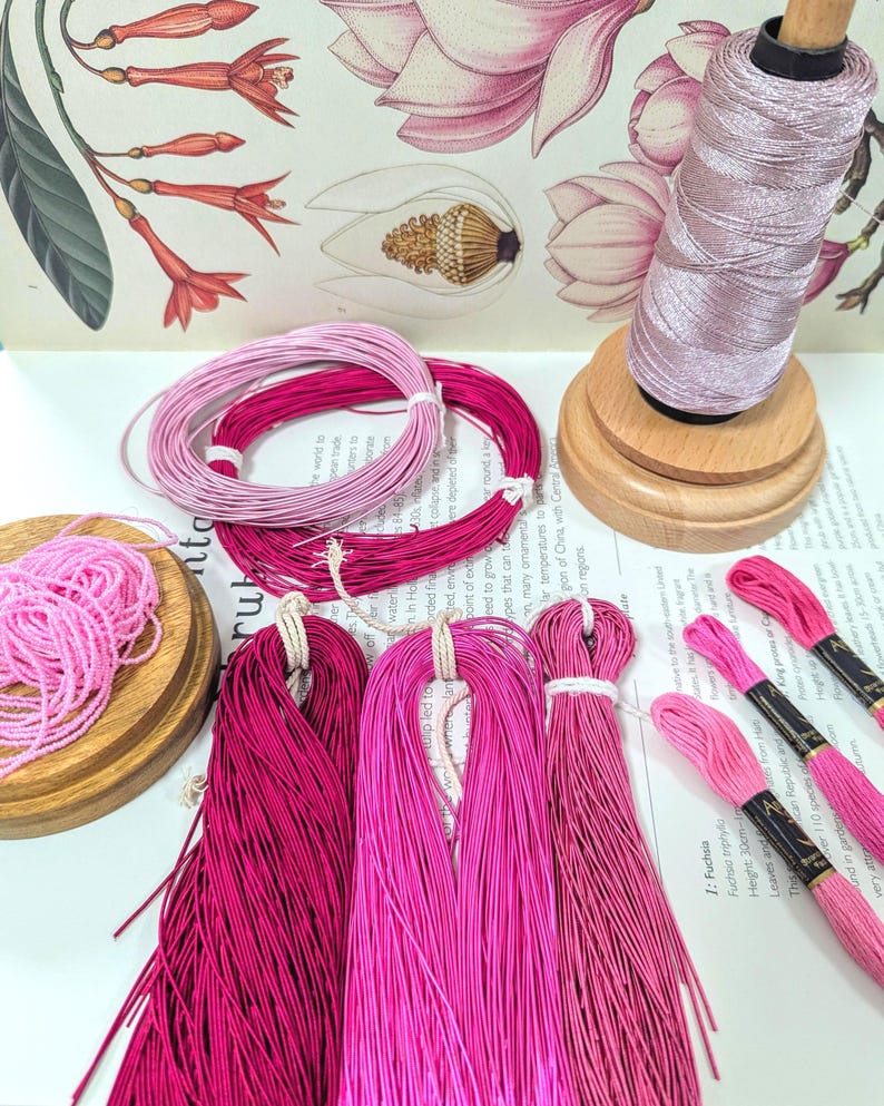 May include: An assortment of pink and fuchsia embroidery floss and thread. Several skeins of floss are bundled together, and a spool of thread sits on a wooden holder. The background features floral illustrations.