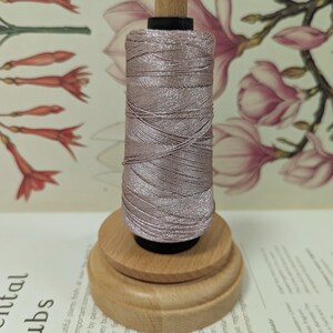 May include: A spool of light mauve thread sits on a wooden base. The thread is tightly wound, and the wooden base has a tiered design. The top of the spool has a small, light-colored wooden knob.
