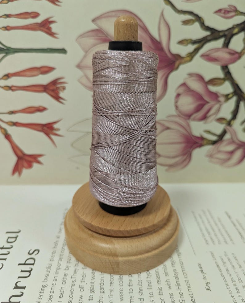 May include: A spool of light mauve thread sits on a wooden base. The thread is tightly wound, and the wooden base has a tiered design. The top of the spool has a small, light-colored wooden knob.