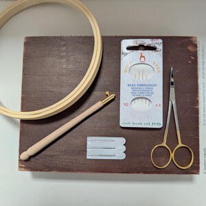 May include: A wooden embroidery hoop, a wooden needle tool, a package of bead embroidery needles, a pair of gold-handled scissors, and a few white needle cards are arranged on a dark brown wooden surface. The text "GOLD EYES" and "BEAD EMBROIDERY" are visible.