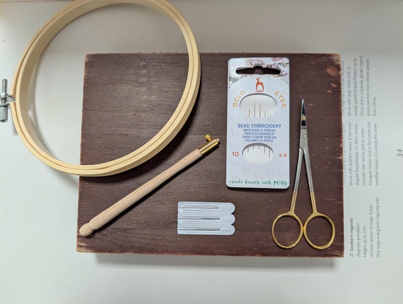 May include: A wooden embroidery hoop, a wooden needle tool, a package of bead embroidery needles, a pair of gold-handled scissors, and a few white needle cards are arranged on a dark brown wooden surface. The text "GOLD EYES" and "BEAD EMBROIDERY" are visible.