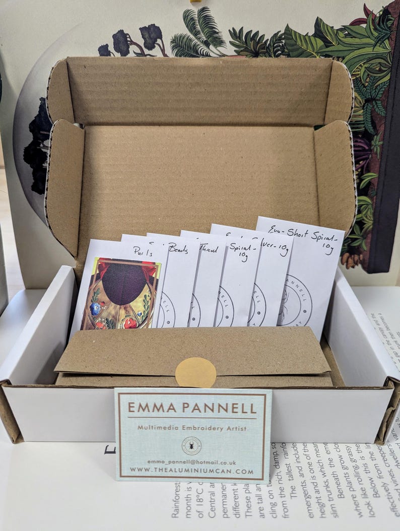 May include: An open cardboard box containing embroidery supplies. Inside are small packets labeled "Parls", "Beads", "Thread", and "Spiral". A business card for Emma Pannell, Multimedia Embroidery Artist, is visible.
