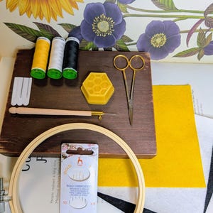 May include: A craft kit with embroidery supplies. Includes a wooden embroidery hoop, needles, thread spools in yellow, white, and black, a wooden needle holder, scissors, a yellow honeycomb-shaped wax, and felt fabric. The kit also includes a package of bead embroidery needles.