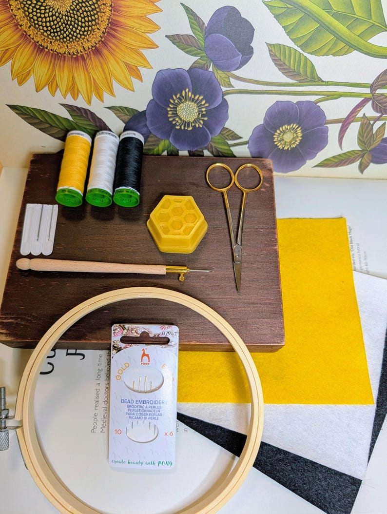 May include: A craft kit with embroidery supplies. Includes a wooden embroidery hoop, needles, thread spools in yellow, white, and black, a wooden needle holder, scissors, a yellow honeycomb-shaped wax, and felt fabric. The kit also includes a package of bead embroidery needles.