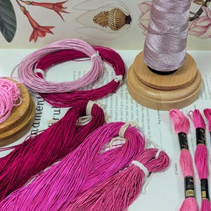 May include: An assortment of pink and magenta embroidery floss and thread. Several skeins of floss are bundled together, and a spool of thread sits on a wooden base. The image includes botanical illustrations in the background.