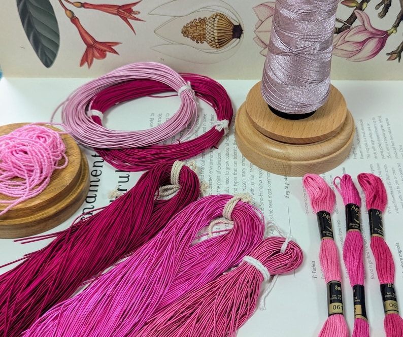 May include: An assortment of pink and magenta embroidery floss and thread. Several skeins of floss are bundled together, and a spool of thread sits on a wooden base. The image includes botanical illustrations in the background.