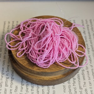 May include: A pile of light pink seed beads on a small, round, wooden base. The beads are strung together, creating a tangled, textured appearance. The wooden base sits on top of a book with printed text.