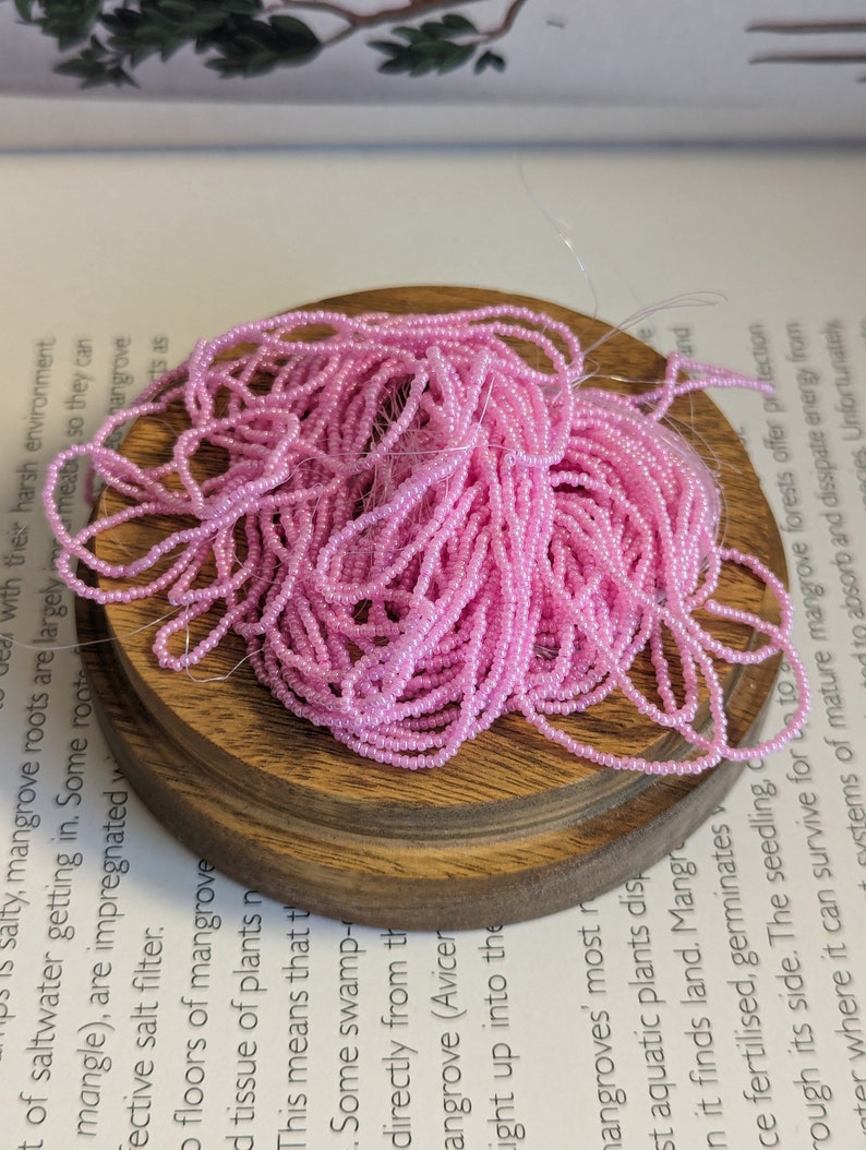 May include: A pile of light pink seed beads on a small, round, wooden base. The beads are strung together, creating a tangled, textured appearance. The wooden base sits on top of a book with printed text.
