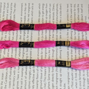 May include: Three spools of bright pink embroidery floss, each with a black label. The floss is neatly wound and ready for crafting. The background is a printed page with text about mangrove forests.