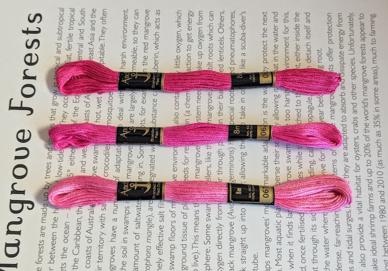 May include: Three spools of bright pink embroidery floss, each with a black label. The floss is neatly wound and ready for crafting. The background is a printed page with text about mangrove forests.