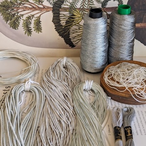 May include: A collection of silver craft materials, including spools of thread, bundles of cord, and a wooden bowl of beads. Two thread spools have black and green caps. The background includes botanical illustrations and text.