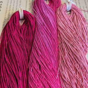May include: Three bundles of pink and magenta wire, each tied with a white cord. The wire colors range from deep magenta to light pink, creating a gradient effect. The wires are tightly packed, suggesting a craft or jewelry-making material.