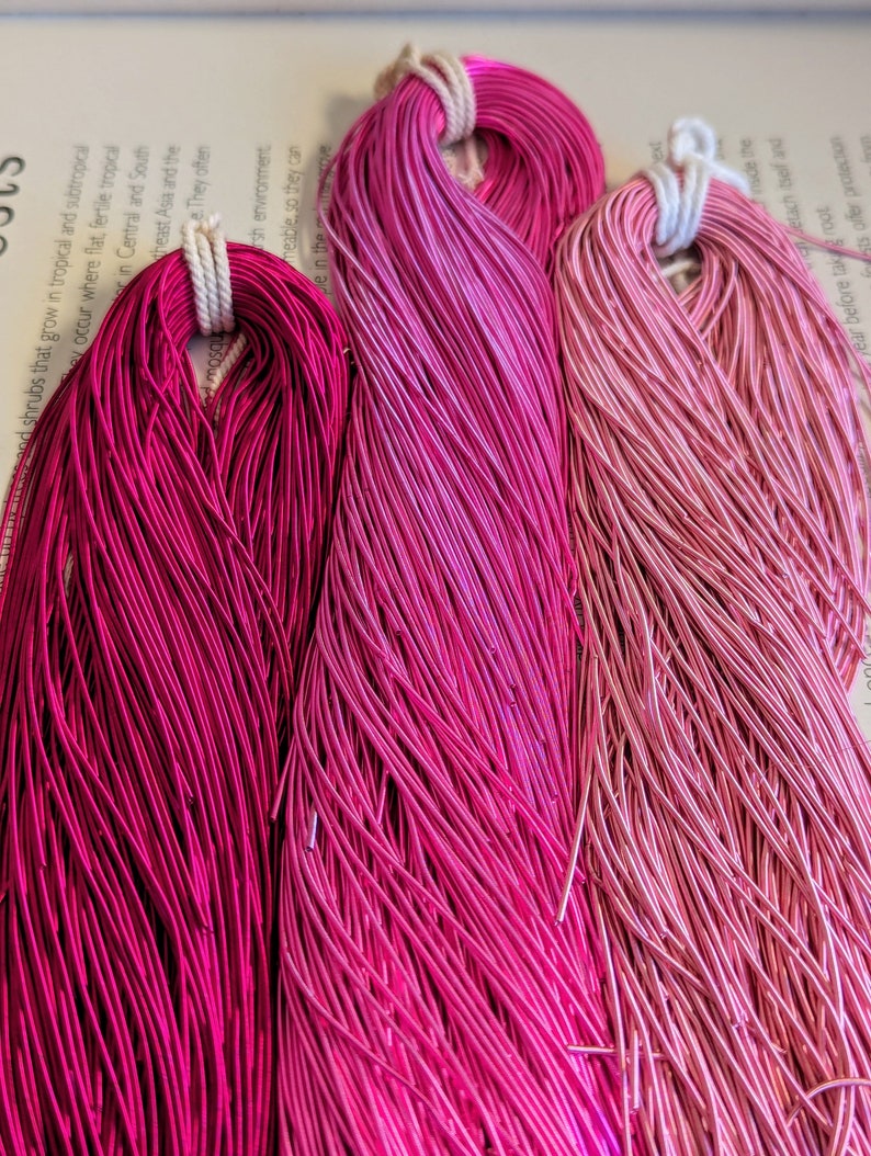 May include: Three bundles of pink and magenta wire, each tied with a white cord. The wire colors range from deep magenta to light pink, creating a gradient effect. The wires are tightly packed, suggesting a craft or jewelry-making material.