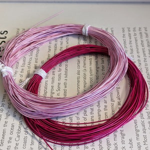 May include: Two spools of wire in shades of pink and magenta, bundled with white string. The wires are coiled on top of an open book with text about mangroves.