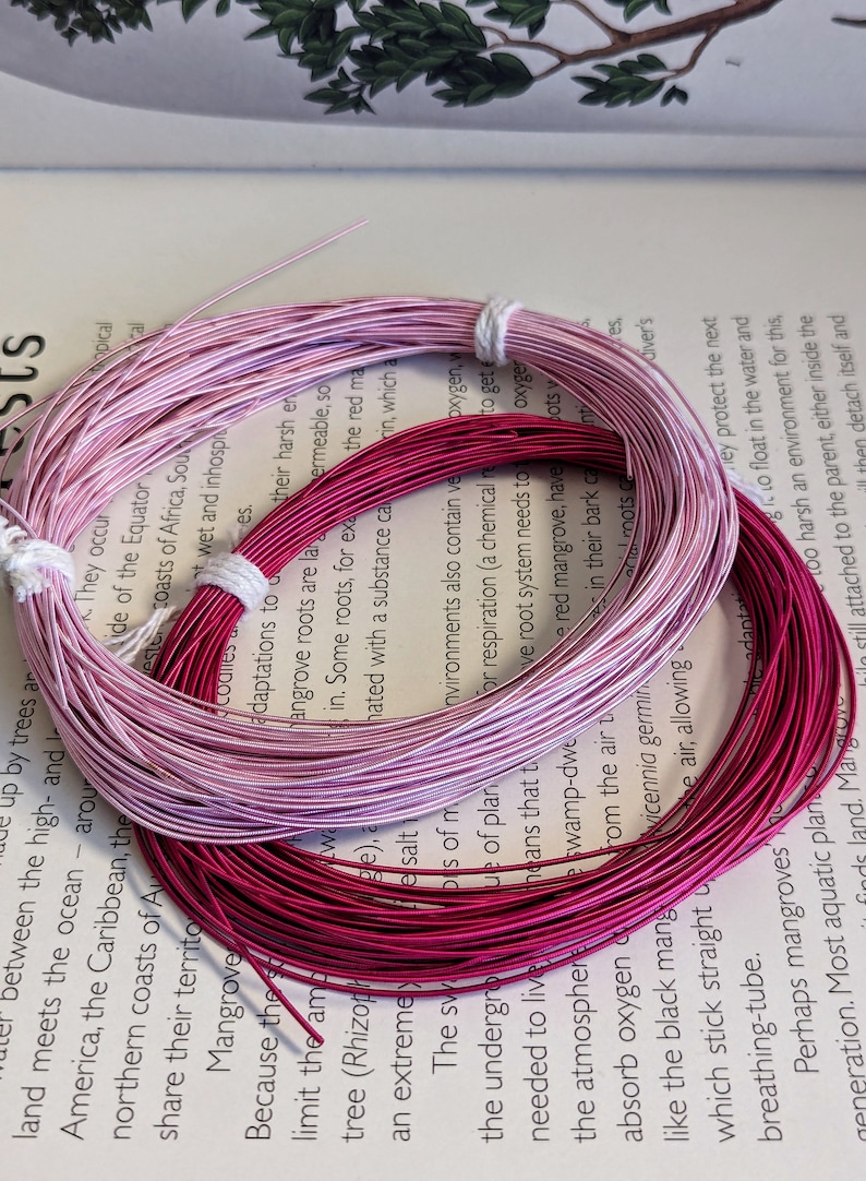 May include: Two spools of wire in shades of pink and magenta, bundled with white string. The wires are coiled on top of an open book with text about mangroves.